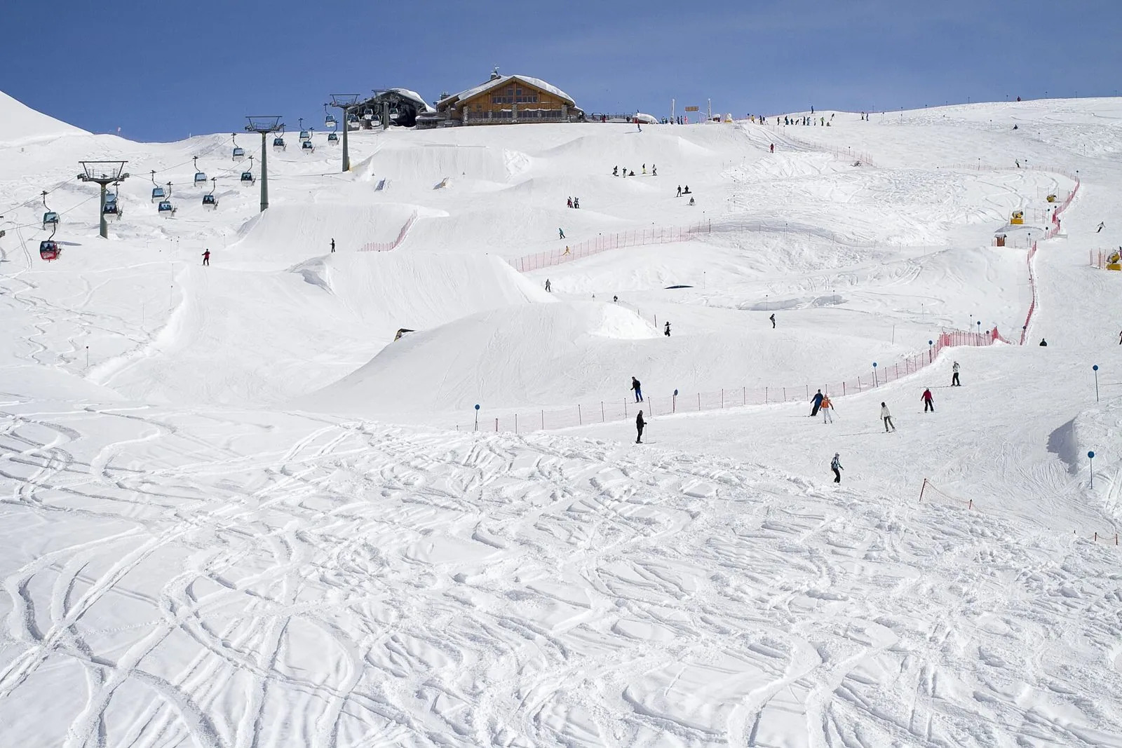 Pista da sci innevata con sciatori lontani, una seggiovia e un rifugio in legno sulla cima, sotto un cielo azzurro